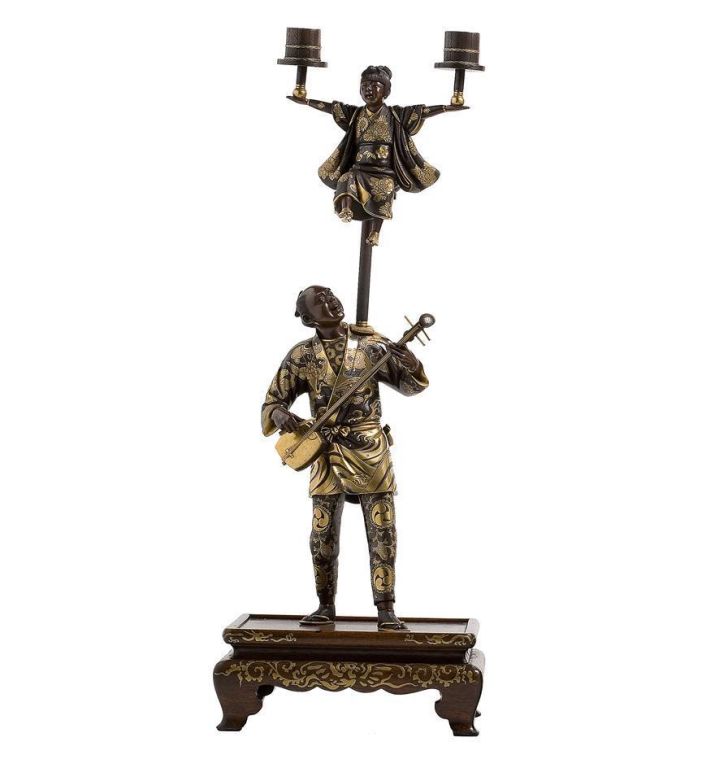 A very fine pair of Japanese multi-patinated bronze candelabra, each a figure of a street entertainer with elaborately inlaid <br />
robes, balancing a boy on his shoulders who holds aloft a pair of candlesticks, on inlaid rosewood stands, signed