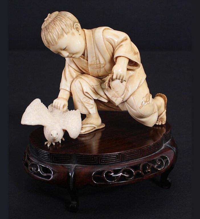A Japanese Meiji period ivory carving of a kneeling boy and bird, on a hardwood stand.