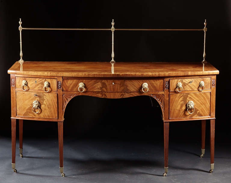 A mahogany George III bowfront sideboard with brass gallery. The inlaid and carved case is supported by square tapered, reeded legs on brass casters. The top with crossbanded and inlaid edge conforms to a bowed front case with a central silver