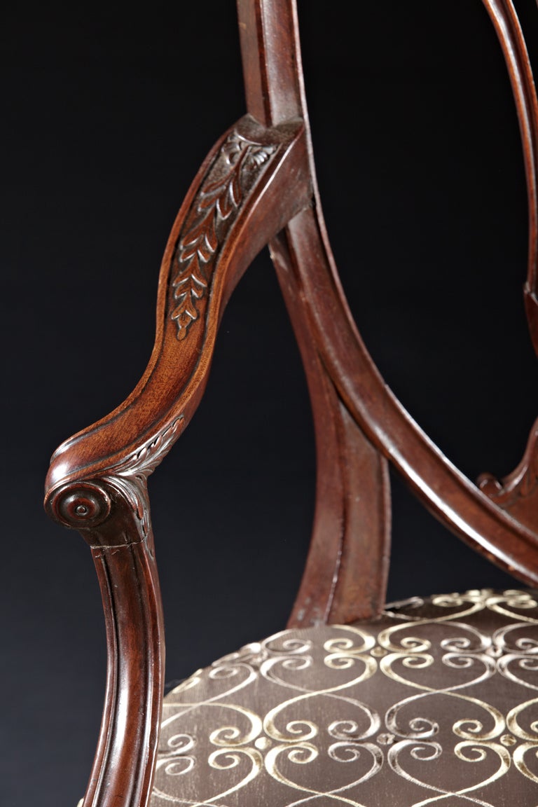  	A pair of mahogany carved Hepplewhite armchairs with shield shaped backs attributed to Gillows. The pierced splats with carved acanthus and rosettes are within a shield shaped frame with similar decoration over upholstered seats with bell-flower