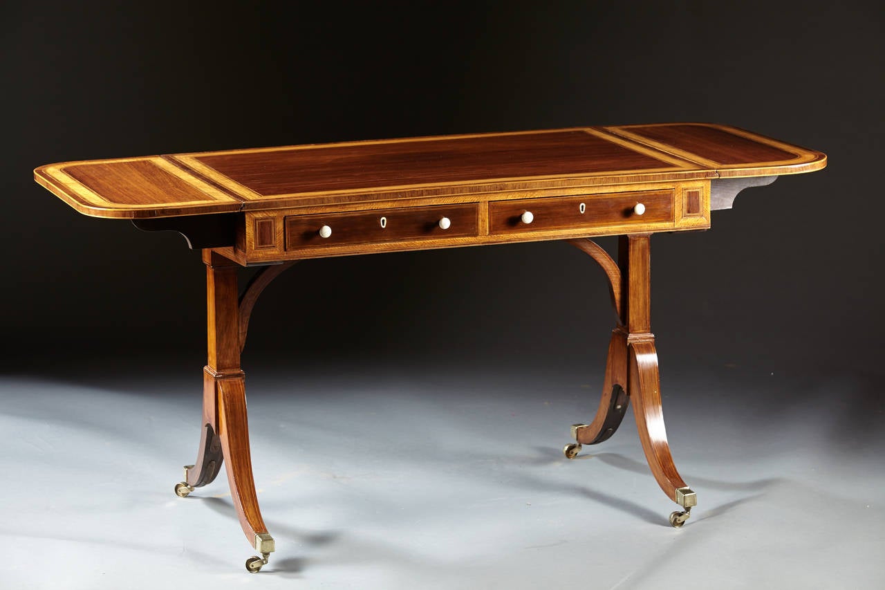 A finely executed English Regency period mahogany sofa table cross banded in wide band satinwood with line inlay. The double drop leaf top above a case with two working drawers to one side and false drawers to the reverse. The case supported by a
