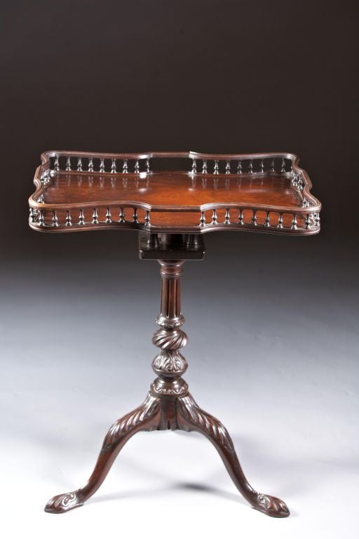 A George II-III mahogany galleried tilt and turn tea table with carved tripod stand. The figural mahogany rectangular shaped top with brass inlaid railing and turned baluster gallery is supported by a birdcage mechanism. The shaft with fluted column