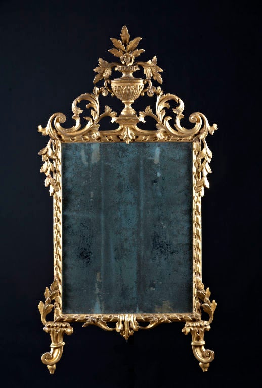 An Italian neo-classic giltwood carved  mirror having a foliate and urn crest above a ribbon carved frame ending in drop carved scroll feet. Italian, circa 1770. Related examples in World Mirrors, Graham Childs, p. 284, pl. 629.