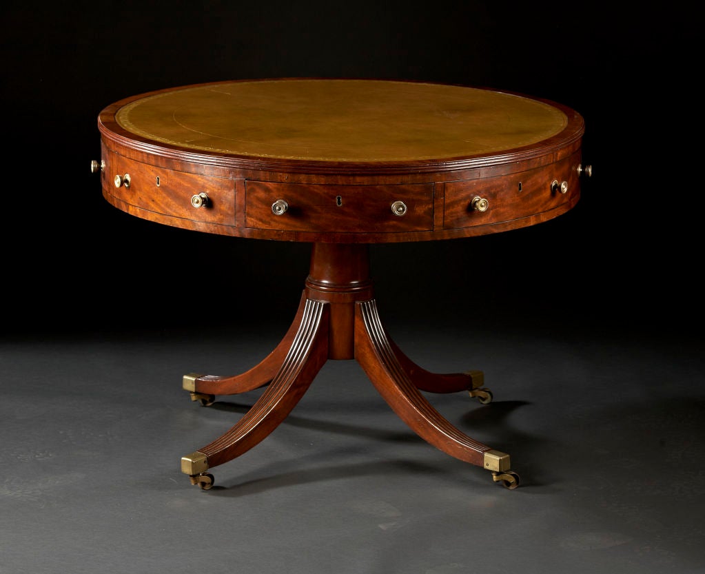 A Sheraton mahogany drum table with green leather top over alternating sham and working drawers in the frieze on a column shaft supported by downswept, fluted legs on brass casters. The drawers are finely lined in cedar. English, circa 1790-1800.