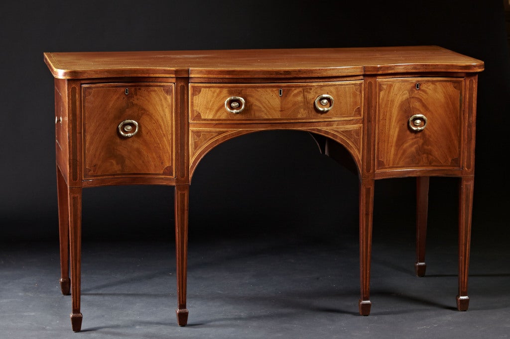 A very fine serpentine sideboard from the early Sheraton period, circa 1790. The mahogany case with three drawers is inlaid throughout with satinwood stringing and harewood crossbanding. The serpentine case is supported by square tapered, inlaid