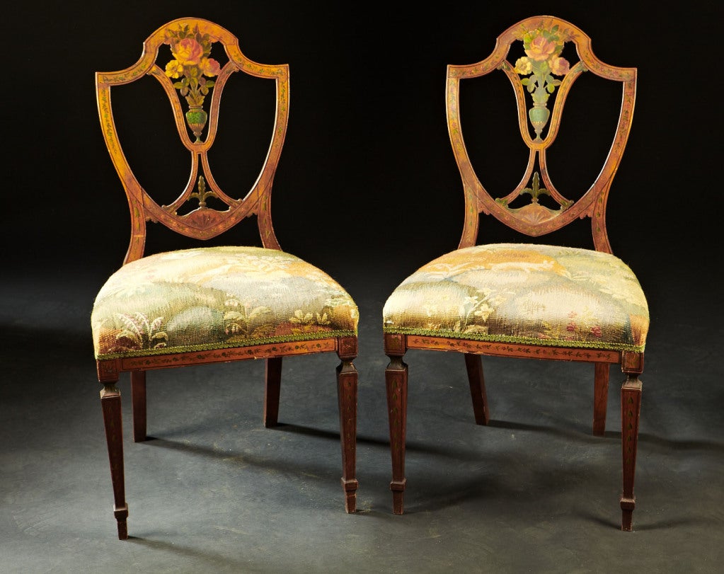 A pair of English, Hepplewhite period, paint decorated satinwood side chairs. The shield shaped backs are decorated with floral and bellflower designs. The legs are square tapered and decorated in the same fashion. English, circa 1770-90.