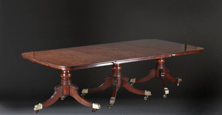 A Regency mahogany three pedestal dining table with figured crotch grained mahogany top on boldly turned shafts with hipped and carved knees above downswept and  reeded legs ending in large, fluted brass castered feet. English, circa 1810. Without