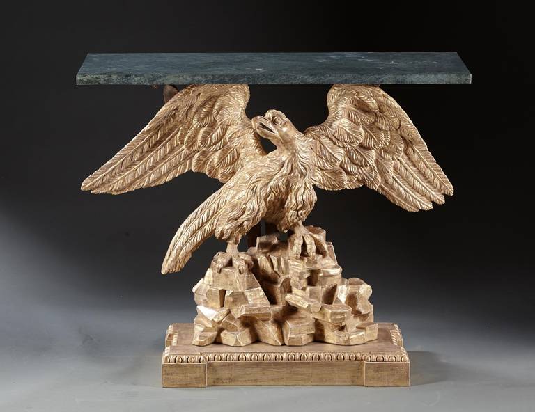 A Regency carved and gilded eagle console table. The majestic spread winged eagle is perched on a rock foundation over an egg and dart carved plinth base. The later green marble top is supported by an iron armiture behind the eagle. 19th century