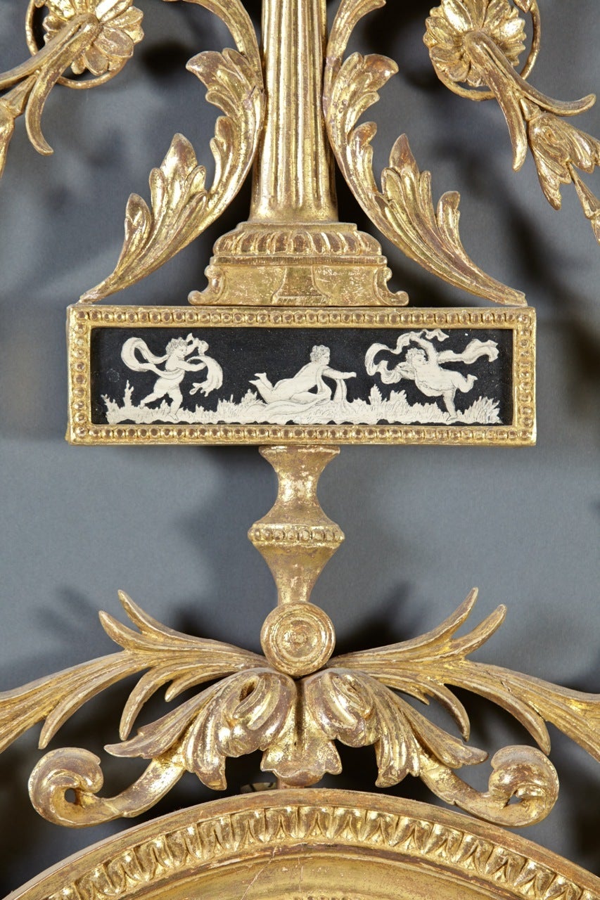A rare English Hepplewhite period giltwood mirror having a trophy crest above a penwork tablet with neoclassical motifs and foliate scrolls over an imbricated oval frame with bellflower pendant decoration, English, circa 1770.