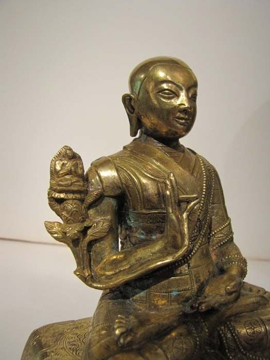 Sinot-Tibetan Gilt Bronze Monk Seated in the Lotus Position on a Carpet Covering Two Books.  The Monk's Left Hand is Shown in Meditation or Dhyana Mudra.  The Monk's Right Hand is Shown in the Teaching or Dharmacakra Mudra. Perched on the Monk's