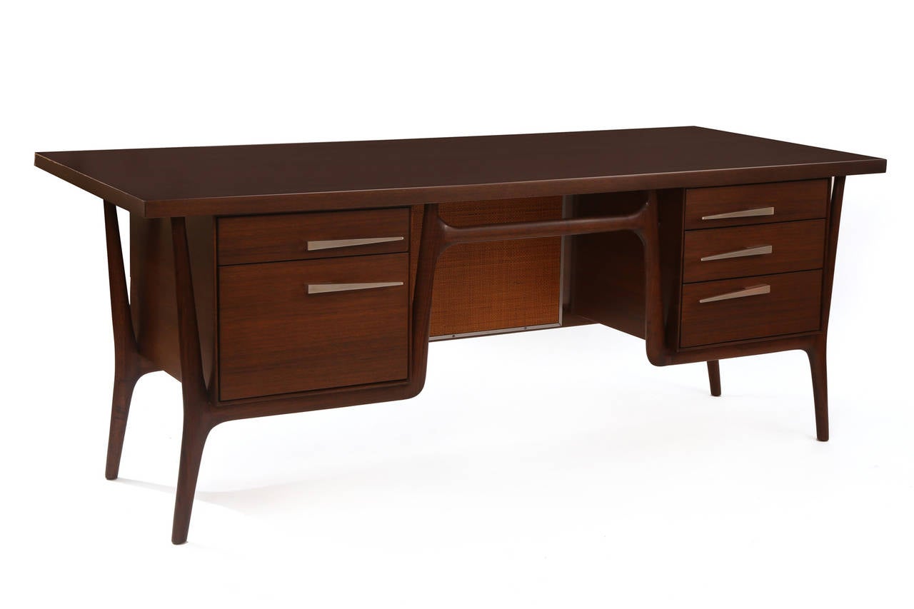 Phenomenal walnut and anodized aluminum desk, circa late 1950s. This example has elegant and sculptural solid walnut legs, anodized aluminum tapered pulls and beautifully grained top. It has five drawers with one file drawer with a wicker and