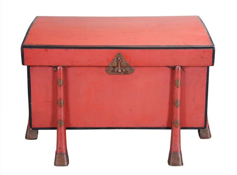 A red lacquered robe chest with a hinged, domed lid. Raised on six legs ending in hoof-shaped feet and embellished with engraved brass details. Outlined with a black lacquered and molded edge.