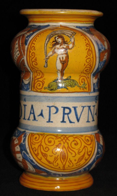 A Castel Durante glazed ceramic albarello, inscribed “DIA. PRVN. SOL” within a central banner below a goddess standing on a stylized dolphin and above openwork scrolls on a ground painted with trophies.