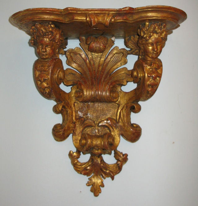 This late Baroque gilded wooden bracket has a shaped top supported by two scrolling herm figures, centering a stylized feather motif above scrolling strap work, and ending in a foliate form.