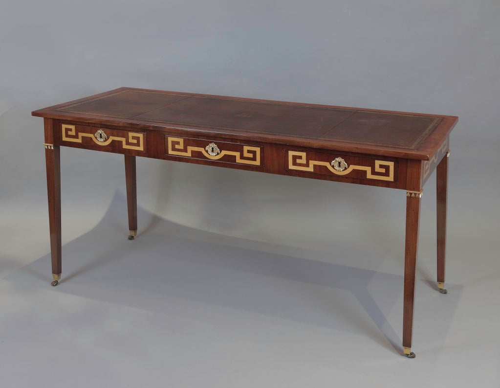 A Neoclassic mahogany desk with three drawers in the front and back; the drawers and sides inlaid in boxwood with a Greek key design, and bronze key escutcheons with acanthus leaf decoration; with tapered legs ending in bronze sabots and rollers.  