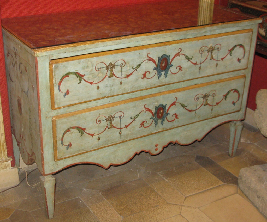 A pale blue painted two-drawer commode with polychrome painted neoclassical motif; the top with faux marble blue and yellow paint; with bronze putti and arrow decorated pulls. The shaped apron with red painted trim.