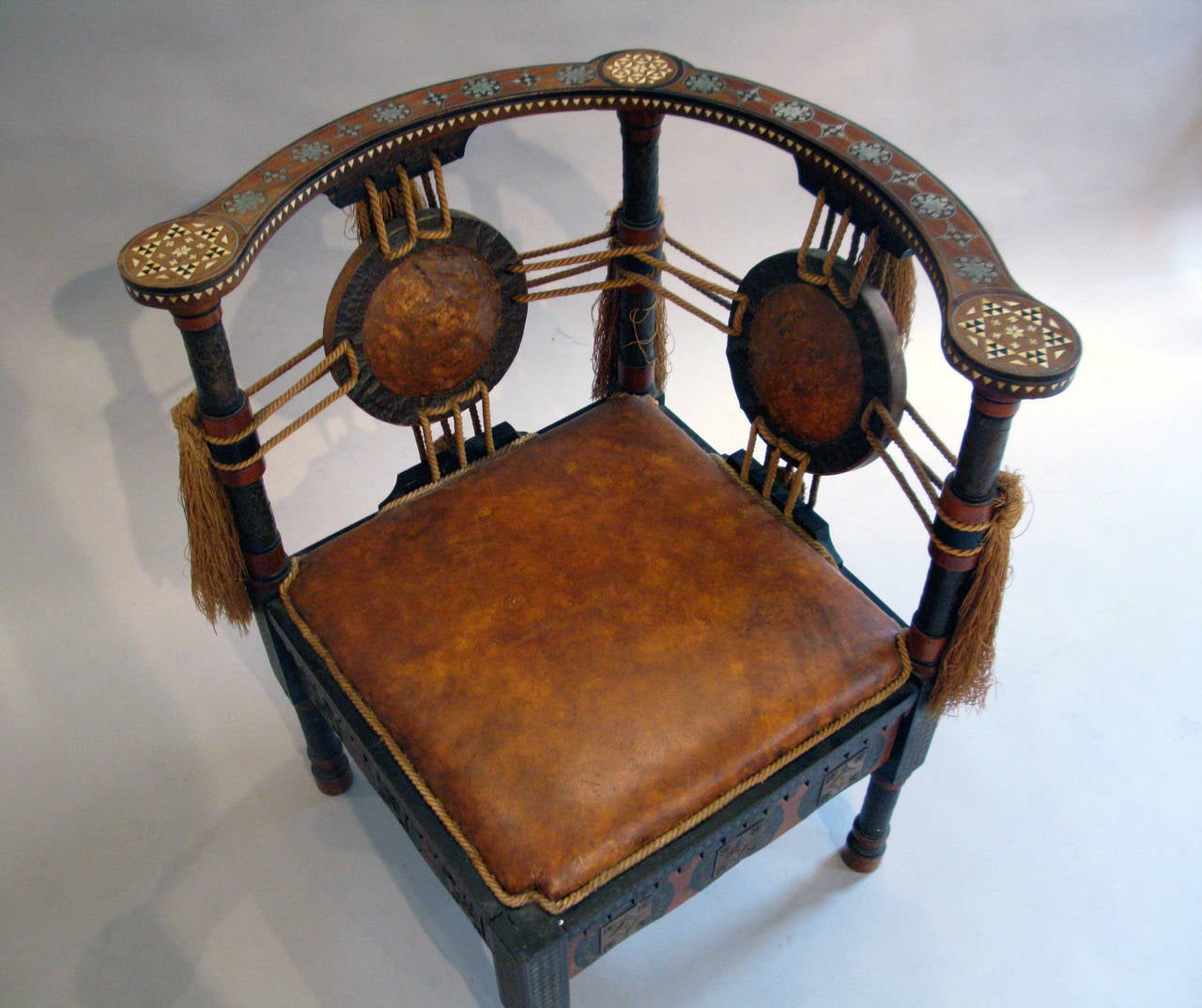 This ebonized walnut corner chair with leather seat and having leather roundels backed by copper panels on turned supports; inlaid with pewter, brass, bone and clad with copper.The roundels supported by silk rope and tassels. In an exotic Moroccan