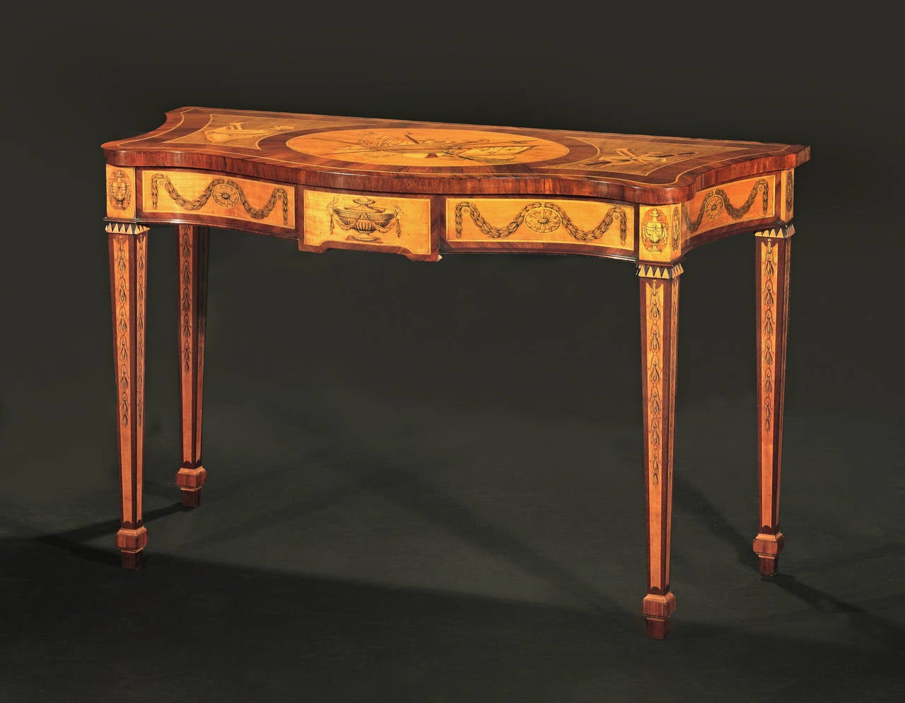 With rosewood, satinwood, harewood and mahogany. The serpentine top having military and musical trophy inlays set within broad banding; the shaped frieze with central classical urn inlaid tablet flanked by superbly executed sunflower roundels hung