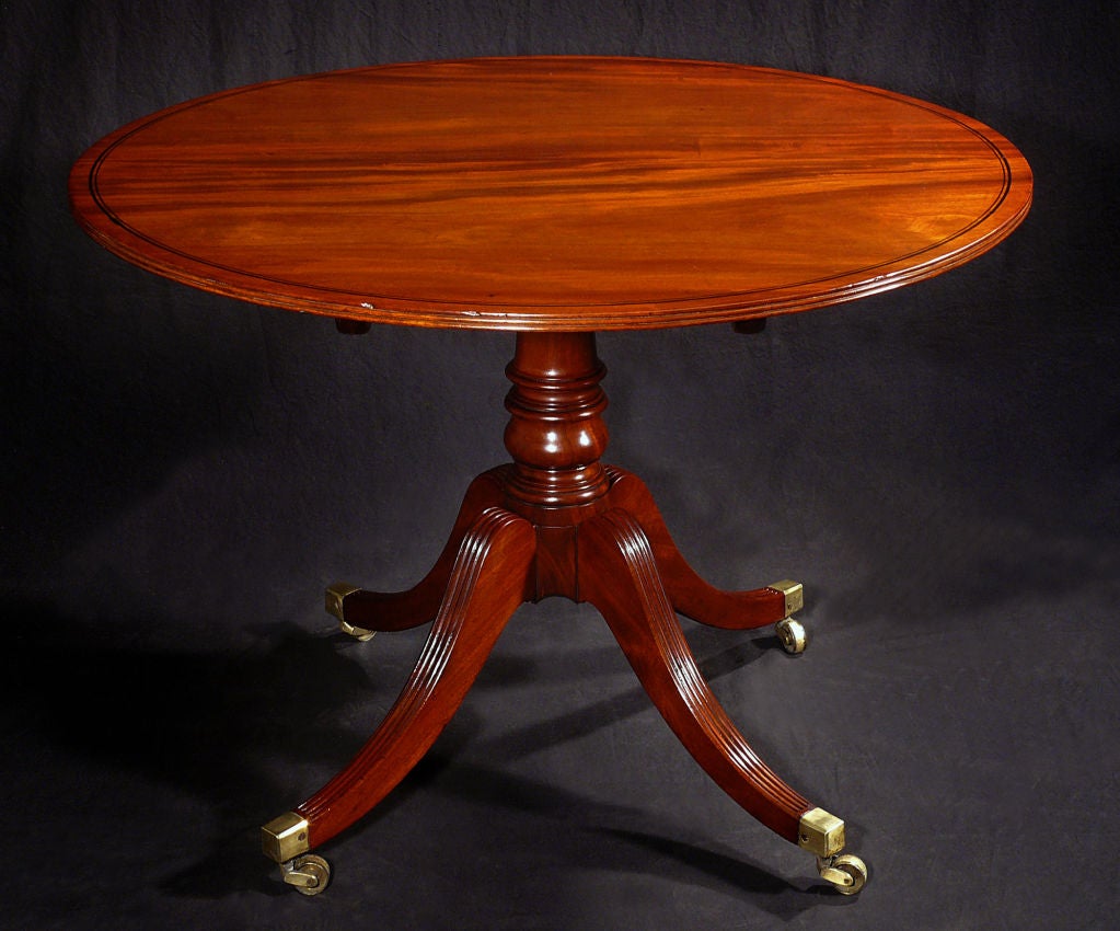 The round tilt-top with ebonized banding over a reeded edge and raised on a ring-turned pedestal support issuing reeded downswept legs ending in brass caps and casters.