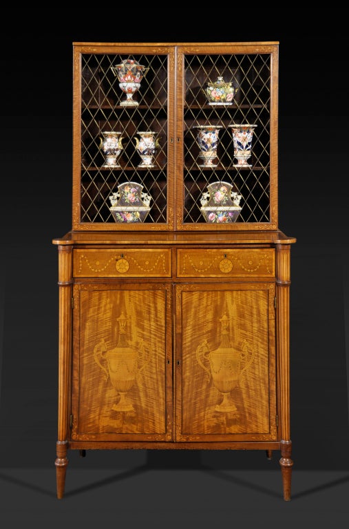 The upper part with a pair of grille-inset doors opening to shelves, over the projecting lower part fitted with two short frieze drawers inlaid with floral roundels and bellflower swags above a pair of Classical urn-inlaid doors flanked by engaged