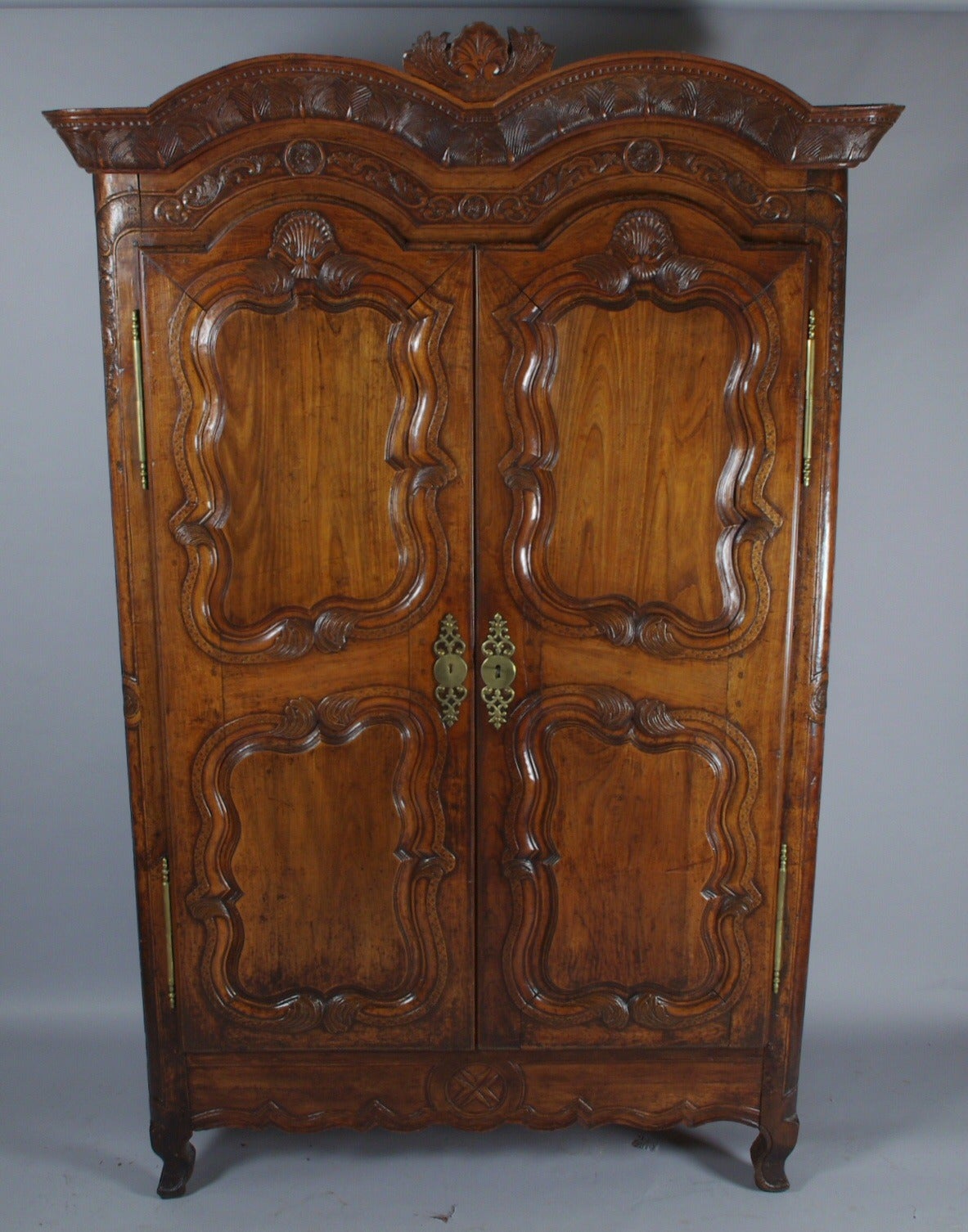Louis XV Provincial walnut double arched armoire, the leaf carved cornice surmounted by a freestanding shell; the two doors, each with inset panels, opening to an interior fitted with modern shelves, on short cabriole legs joined by a shaped apron