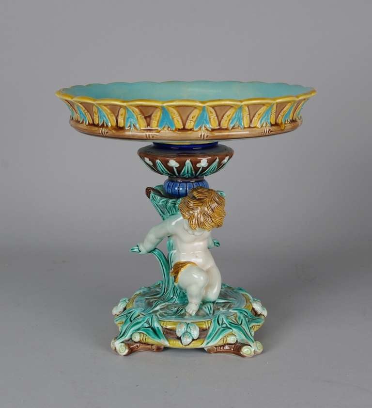 Wonderful Wedgwood majolica compote, the circular turquoise dish with bamboo and leaf border on a flat urn supported by a kneeling cherub (putto) amongst reeds and cattails; the round base with berries, leaves and twigs. Base impressed 