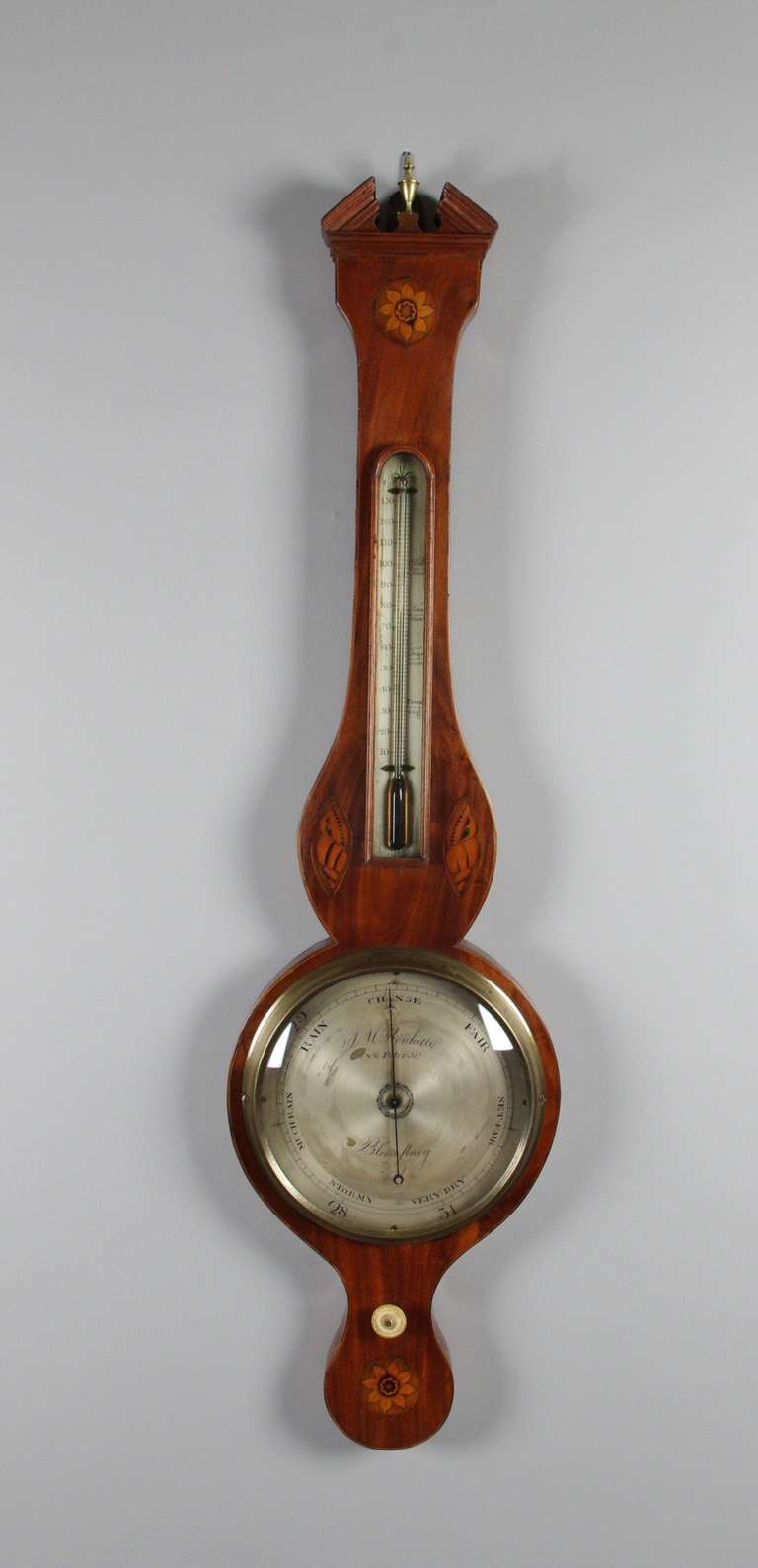 George III mahogany wheel barometer, the broken pediment with a replaced brass finial; the inlaid flower over the thermometer, flanked by inlaid shells; the engraved face with replaced convex glass over the bone set-dial over another inlaid flower.