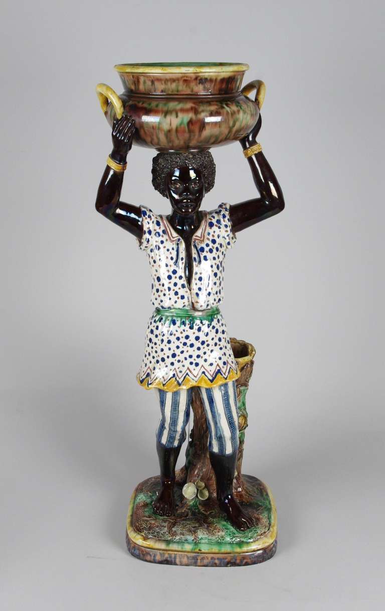 Majolica Blackamoor, the youthful figure dressed in striped leggings and a spotted tunic gathered with a green sash, standing contrapposto near a tree stump and holding on his head an agate glazed urn with yellow handles. 

This wonderful figure