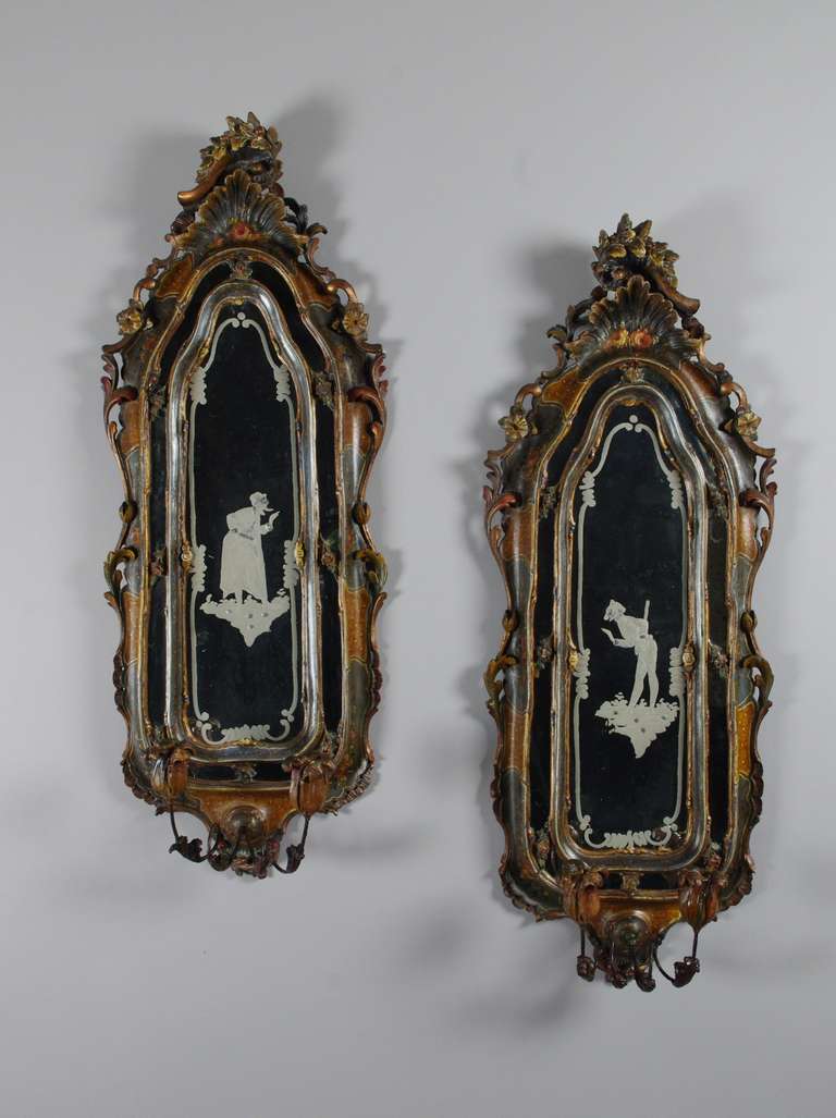 Fine pair of carved and painted girandoles, each with an etched mirror depicting a character from the Commedia dell'arte; the crested frame with a carved shell and trailing leaves, flowers and C-scrolls; the surrounding narrow border of  mirror