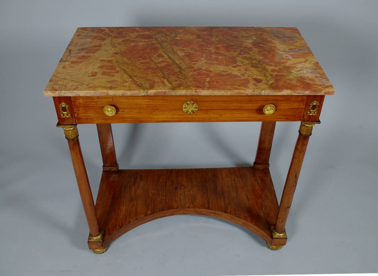 Italian Empire cherrywood console table, the highly figured red marble top (pietra topografica) over a single long drawer with brass knobs centred by a medallion; the turned columns with carved and gilt leaf capitals headed by applied lyres; the