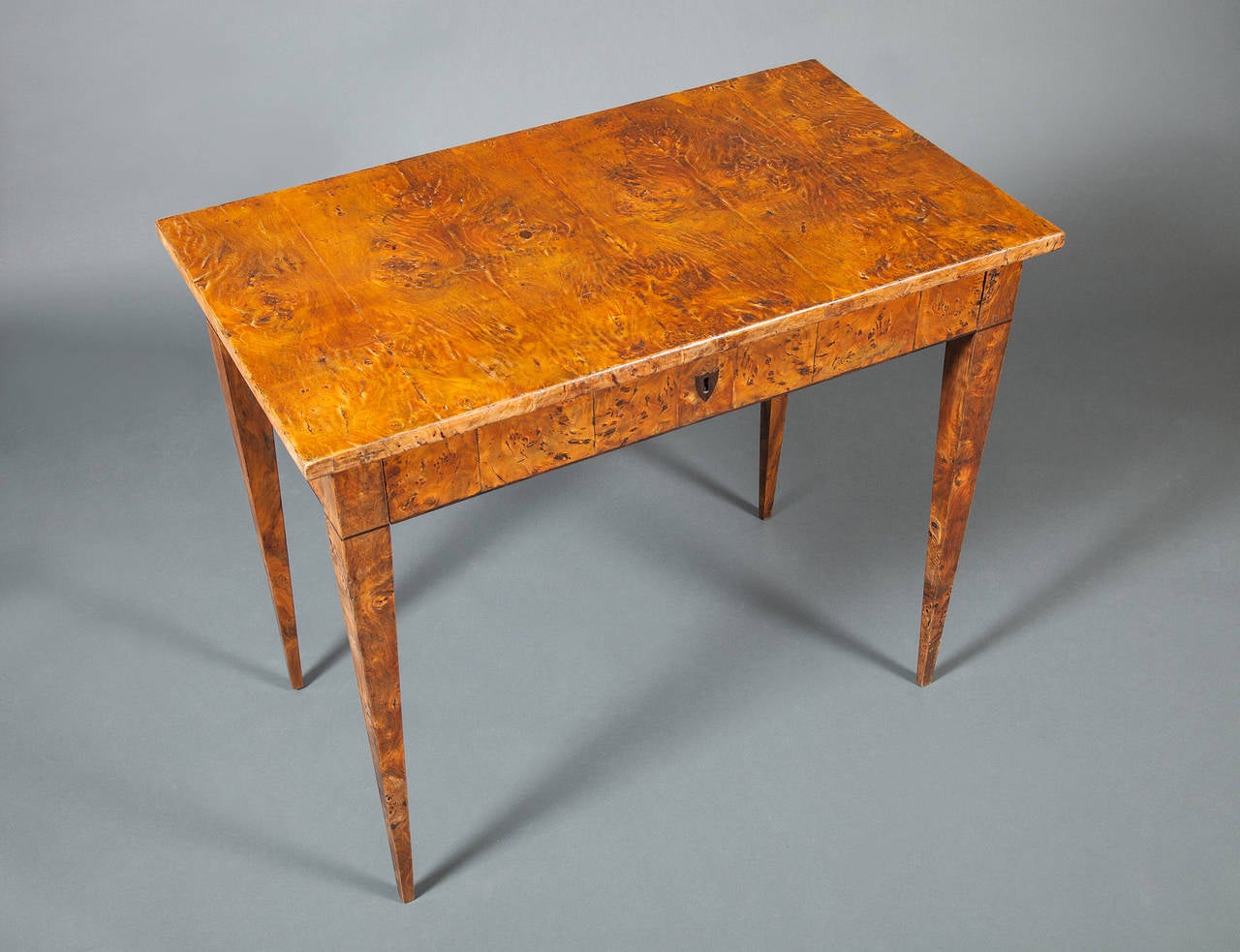Biedermeier burl elmwood writing table. The rectangular over-hanging top above a frieze drawer, fitted with a shield-shaped keyhole escutcheon, raised on square tapering legs, headed by string banding.