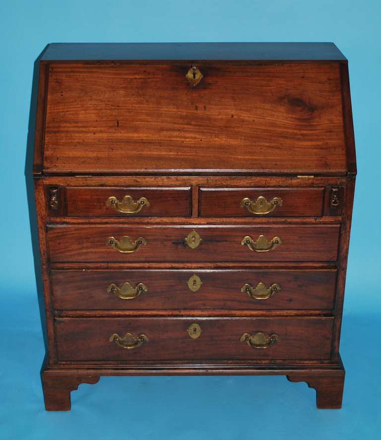 A small George III mahogany slant front bureau, the hinged rectangular top opening to reveal a fitted interior consisting of twin inner and outer compartments of 4 small wells centering a door with brass handle over an arrangement of 2 short side by