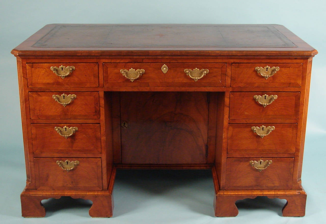 An English walnut pedestal desk with reeded corners, the shaped tooled leather top over nine cross-banded drawers, each with engraved brass batwing pulls, centered by an inset cabinet, resting on bracket feet, circa 1890-1910.