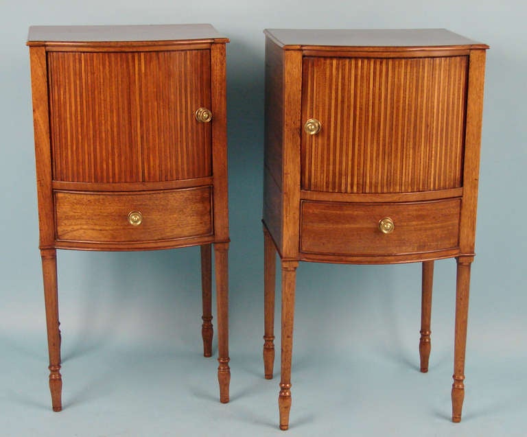 A  rare true pair of Sheraton period mahogany bedside cabinets, each rectangular top with bow front over a tambour door enclosing a later silk-upholstered interior over a silk-lined short drawer; raised on turned legs. Circa 1800.