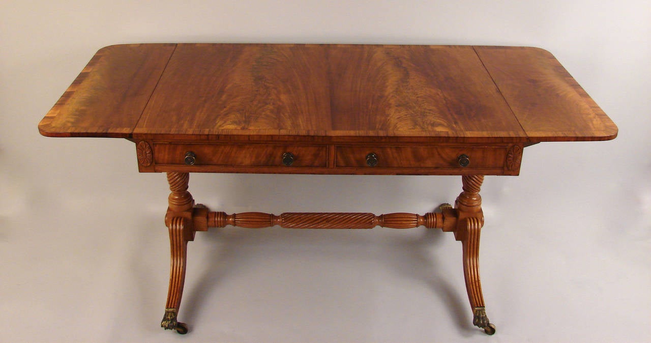 A good quality Regency mahogany sofa table, the well-figured and matched mahogany top crossbanded in rosewood, above two drawers flanked by floral paterae corners, supported on reeded downswept legs connected by a carved and turned stretcher ending