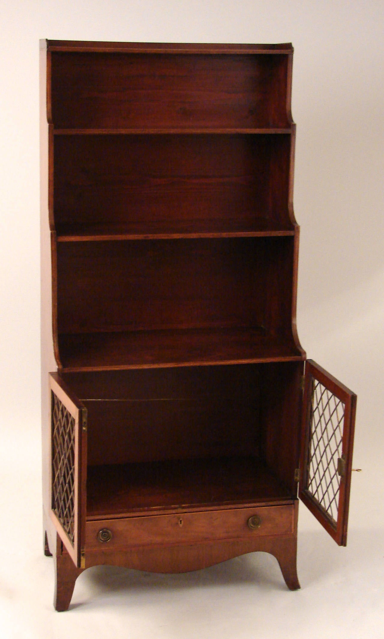 A Regency mahogany and pine waterfall bookcase with three graduated and shaped shelves above a cabinet with brass grill over a single drawer on French feet, circa 1820.