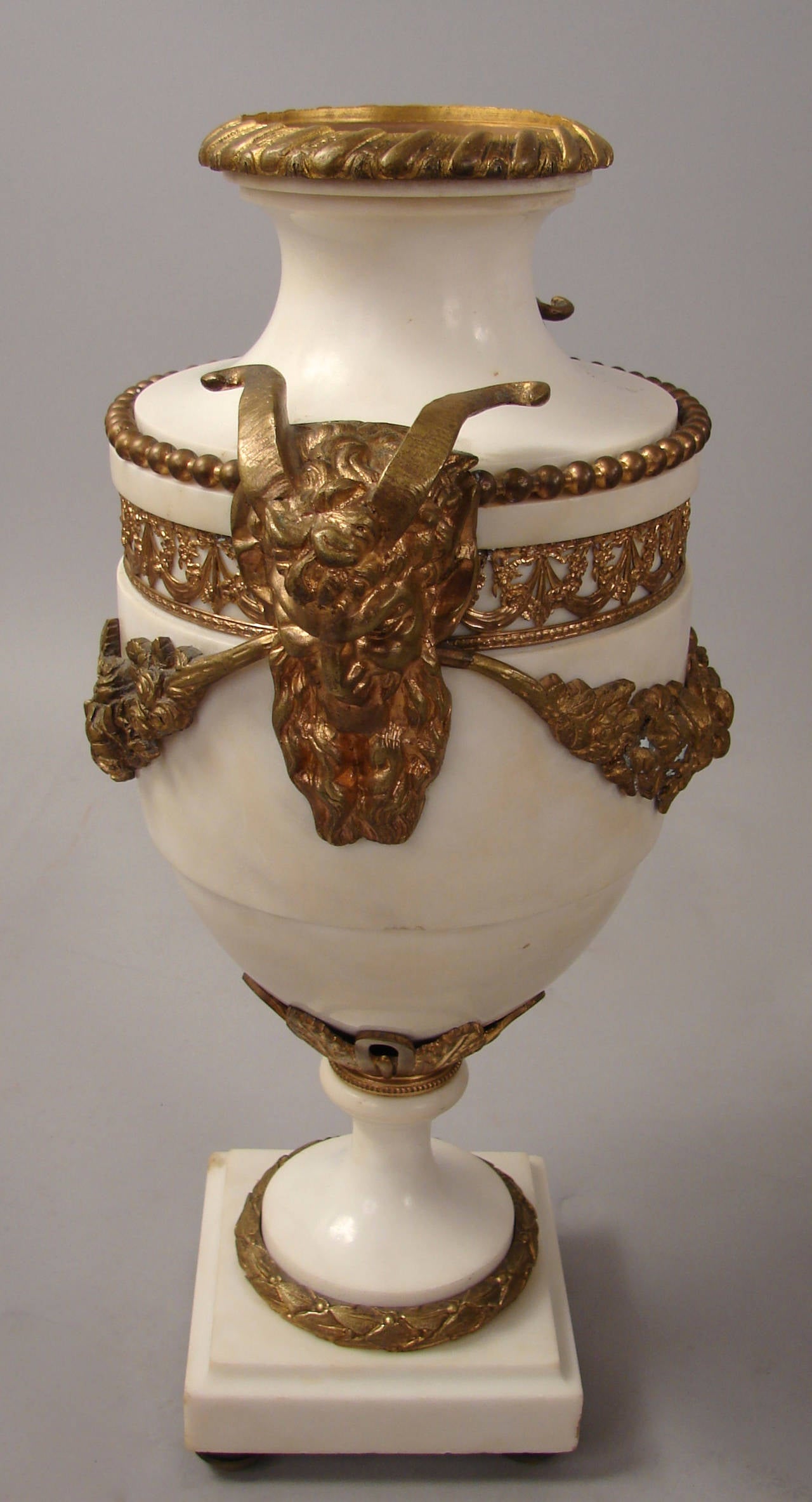 A pair of Louis XVI French neoclassical style gilt bronze and marble urns with rams head mounts above floral swags and rising on a conforming base, circa 1880.