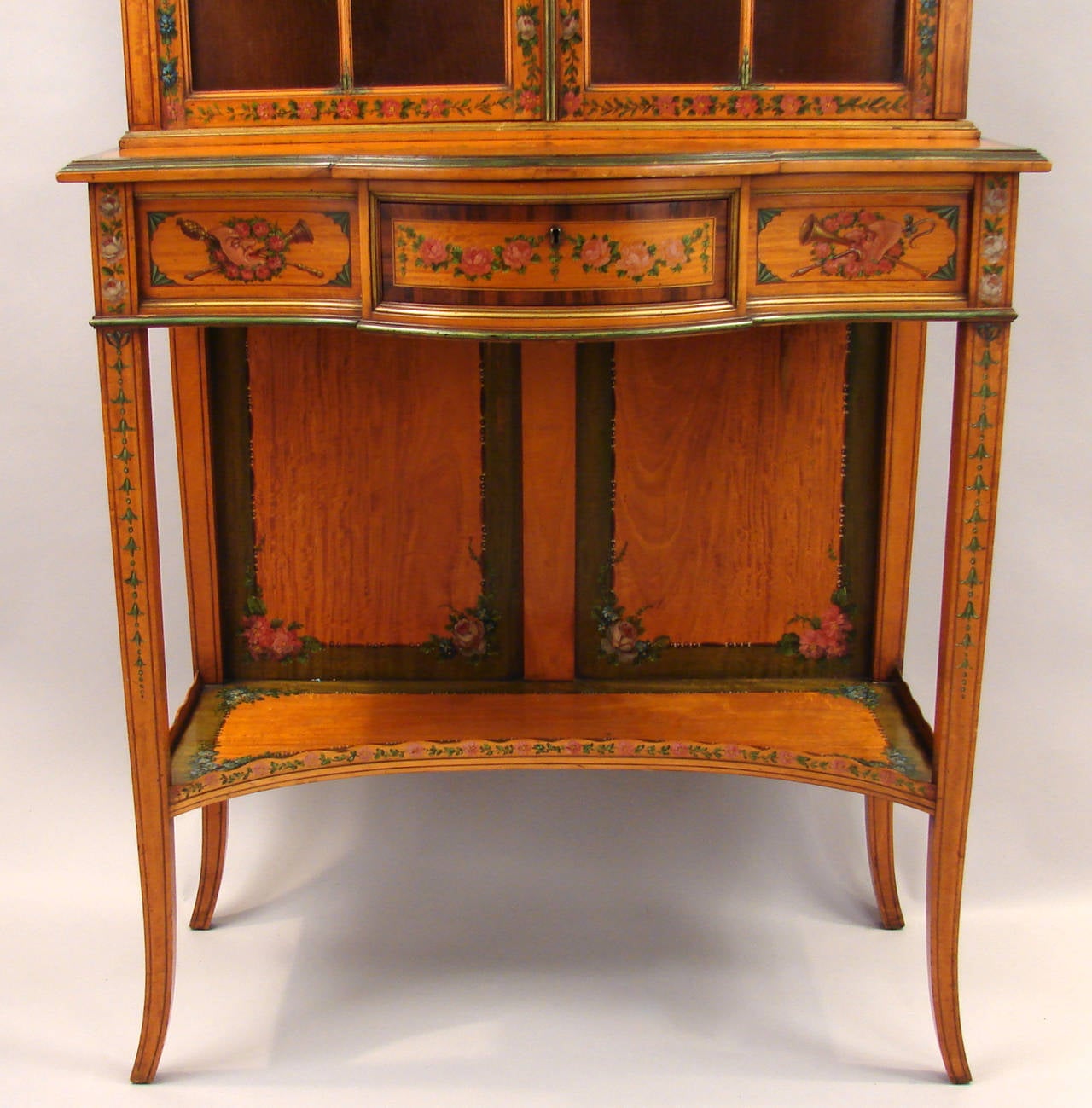 A fine quality English Neoclasssical style paint-decorated satinwood bookcase on stand, having a broken arch pediment with trompe l'oeil dentil molding above astragal glazed doors with elaborate painted decoration in the manner of Angelica Kaufman,