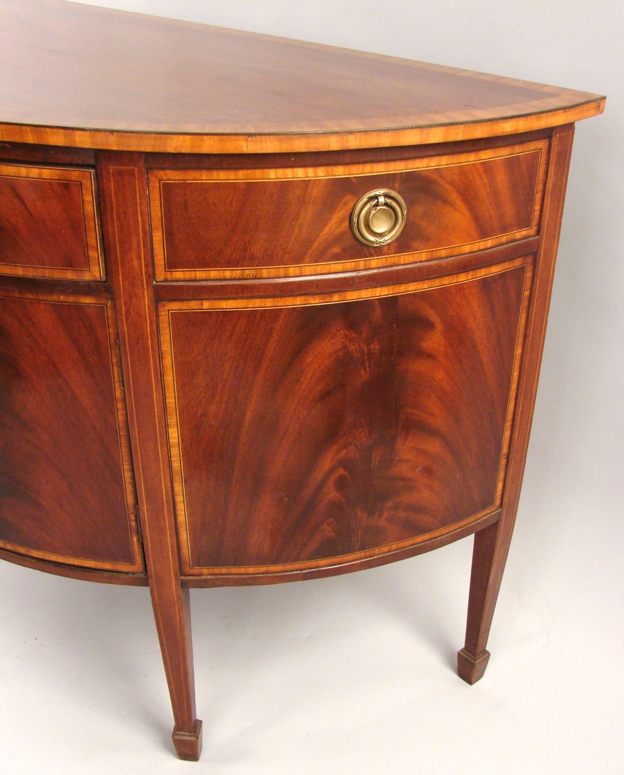 An English Hepplewhite style satinwood inlaid mahogany demilune cabinet, the cross-banded top above one long functional drawer flanked by two false drawers over  figured mahogany cupboard doors, supported by square tapered  legs ending in block