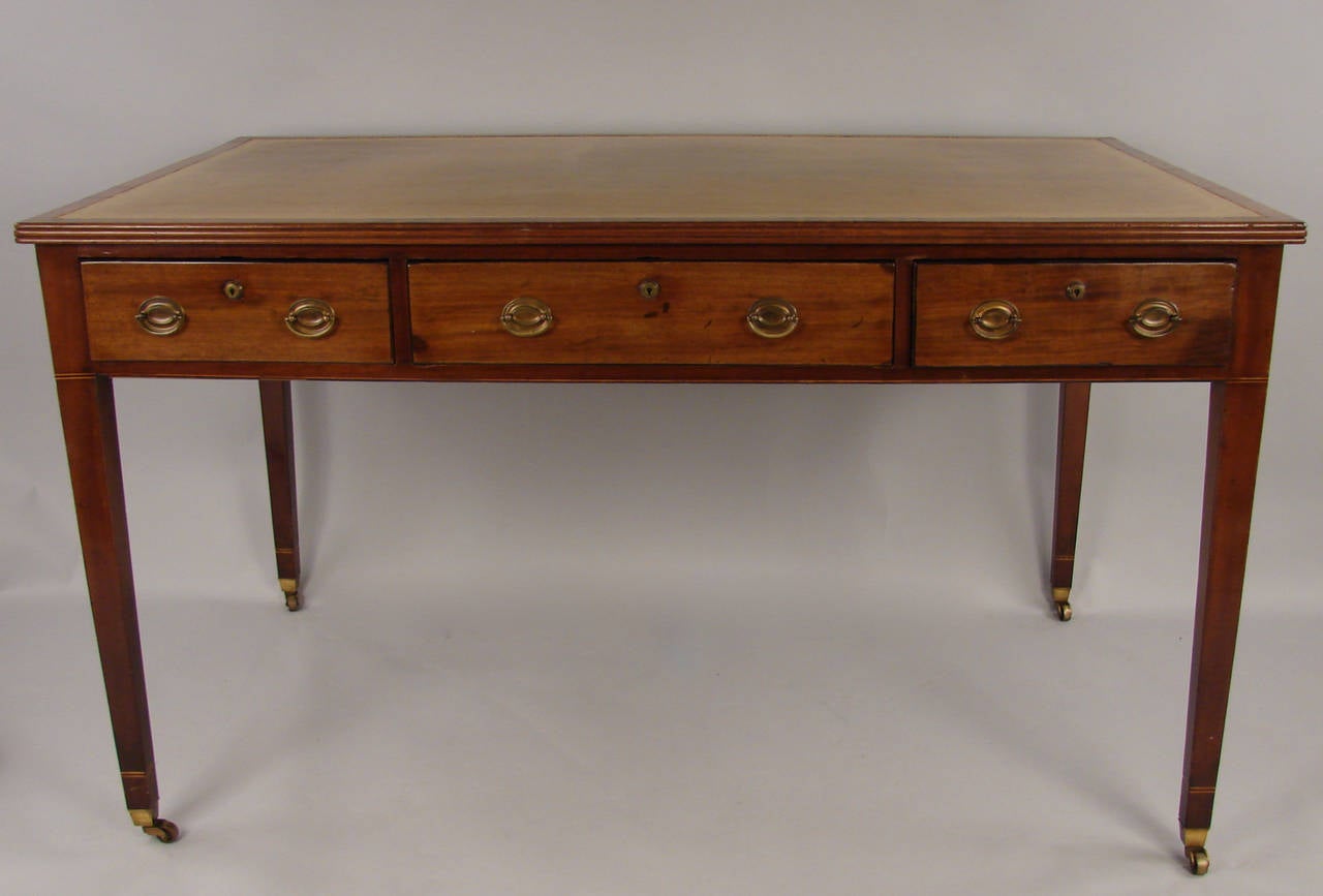 An English inlaid mahogany Hepplewhite period writing table, the tan gilt-tooled inset leather top over three functional frieze drawers, the back decorated with false drawers, the case with line inlay, all on square tapered legs ending in casters.