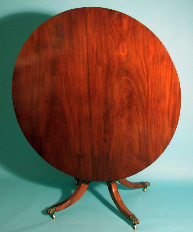 An English Regency mahogany breakfast table, the round tilt-top raised on a turned support and down swept legs ending in gilt paw feet on casters, circa 1825.