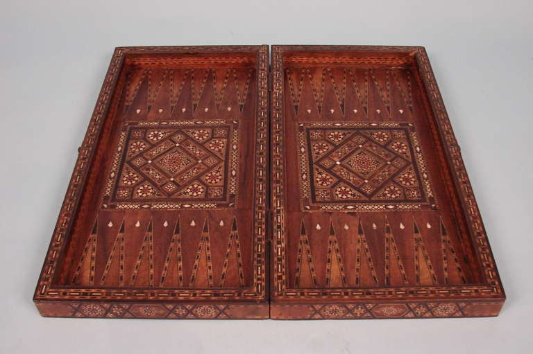 An early 20th century colonial Middle Eastern or North African backgammon set with pieces, elaborately inlaid overall with mother-of-pearl and exotic wood in geometric patterns, the sides with French and Arabic inscriptions. All gaming pieces not