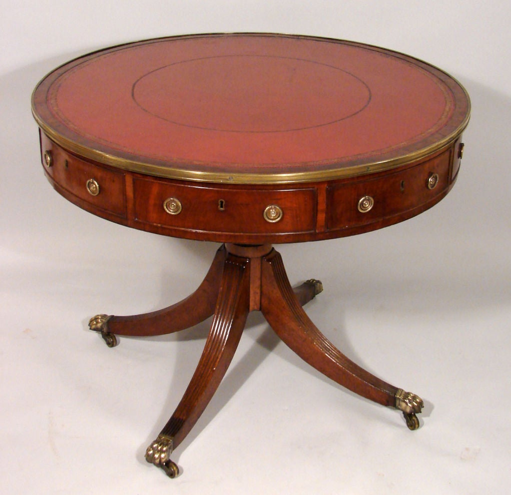 A George III mahogany brass mounted drum table, the circular revolving top inset with red tooled leather fitted with four functional and four false frieze drawers on a turned tapered column and reeded quadripartite supports ending in brass lion paw