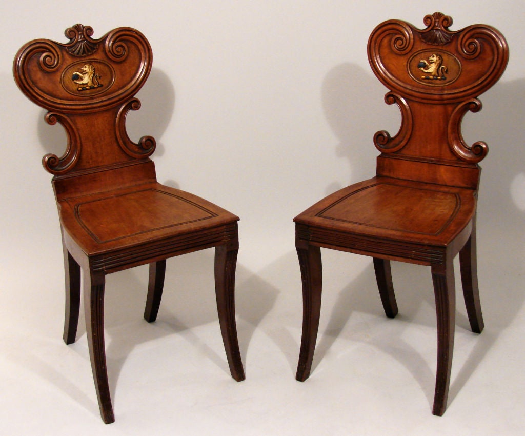 A pair of Regency mahogany hall chairs, each with c-scroll seat back headed by a shell, centering a later painted armorial figure of a lion perched on a barber pole holding a purse, over a trapezoidal seat raised on sabre legs.