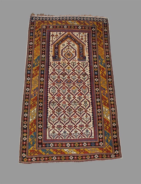 This elegant south Caucasian prayer rug has Classic white field with a variety of flowering plants within a lattice. The 'mihrab' niche is boldly drawn and complimented with a central diamond-shaped form creating what almost looks like a face. The