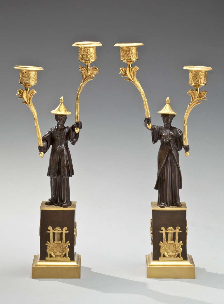 A pair of Empire bronze and ormolu two branch candelabra, taking the form of standing bronze chinese figures (one male, one female) bearing in each hand an ormolu branch with palm leaf torches, on a square black marble plinth with neoclassical