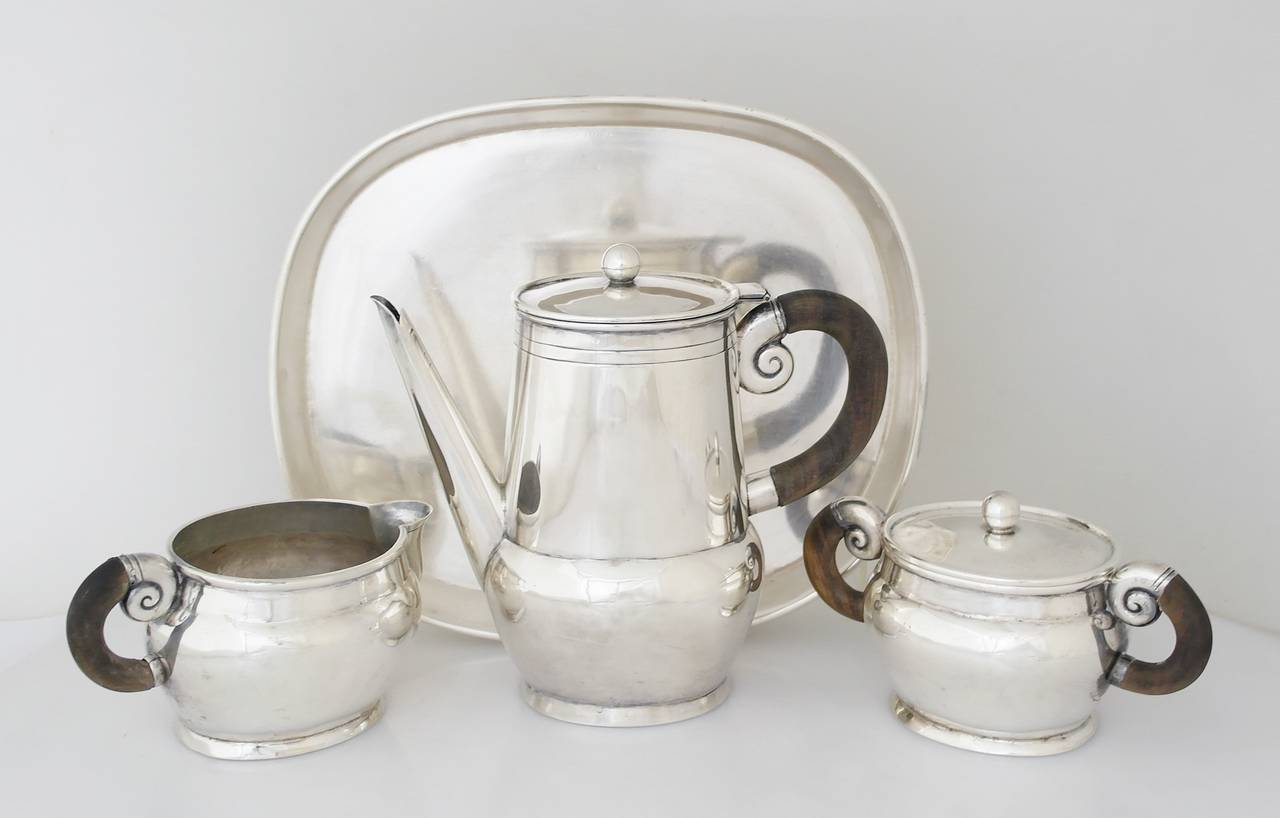 Being offered is a sterling silver tea service by William Spratling of Taxco, Mexico. Comprising a teapot, sugar and creamer all hand-wrought, pot decorated with incised bands; carved rosewood handles; conch motif on handles; matching tray included.
