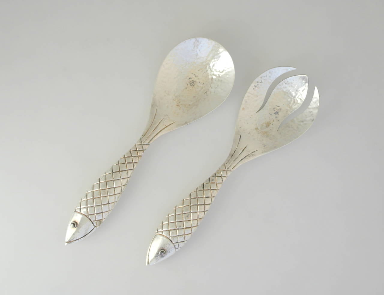 Being offered are a pair of salad servers by Emilia Castillo of Taxco, Mexico. Handmade set with figural fish handles bearing elaborate detail in the scales and eyes, their tails consisting of incised lines leading to the wide bowl and tines, both