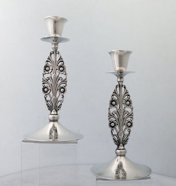 Being offered are a rare pair of sterling silver Tiffany candlesticks, each with a pierced cast solid floriform standard issuing stylized blossoms and leaves, surpounted by a shallow circular drip cup and inverted bell-form candle socket, on a