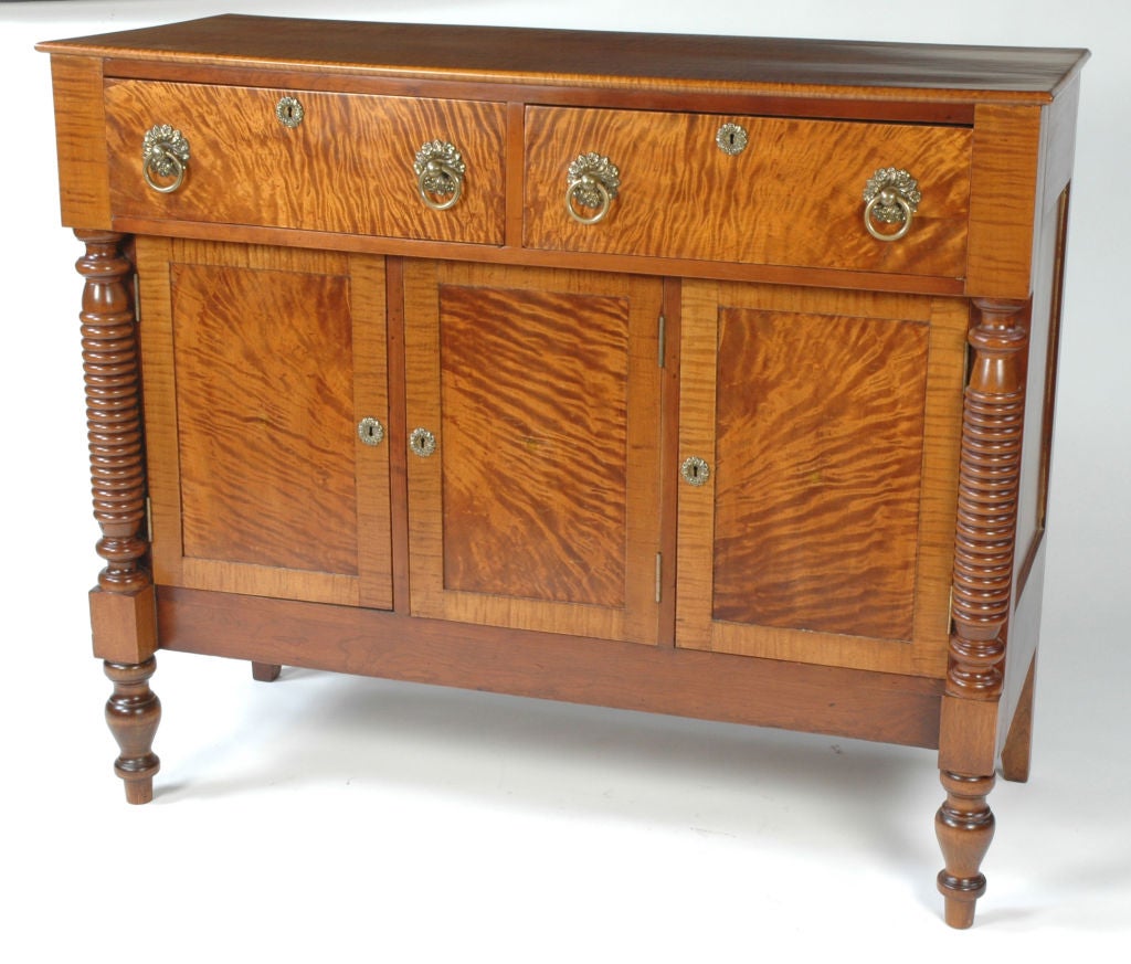 American Sheraton curly maple sideboard/server. The divided top drawer is fitted with its original drop ring brass drawer pulls. The drawer projects over three paneled doors and is supported by flanking turned columns which resolve  into block and